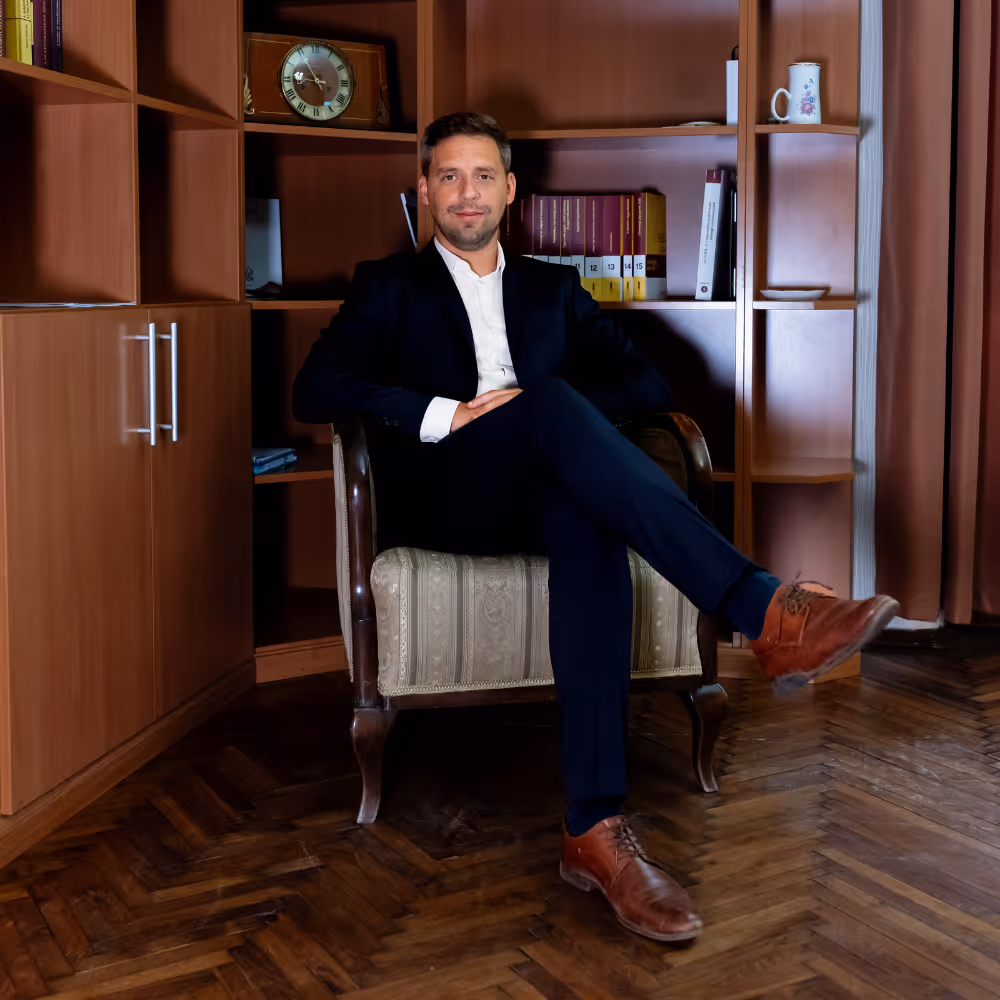 Man in dark suit sitting on an armchair in front of wooden bookshelves with books and clock.